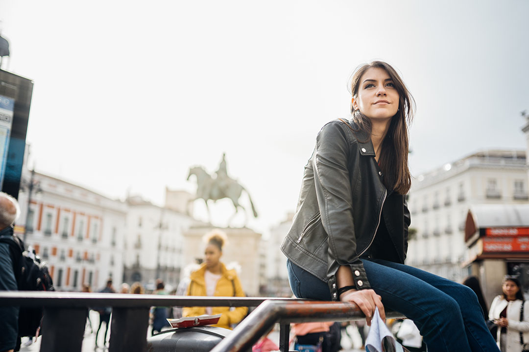 A young woman sitting on a railing with a European statue and plaza in the background